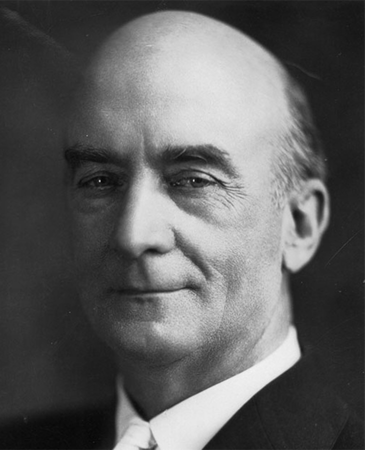 A black and white photograph of a man wearing a suit and tie, with a bald head and visible ears.