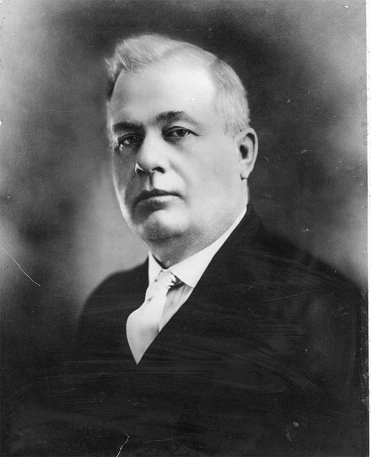 A black and white photograph of a person wearing a suit and tie, centered against a plain dark background.