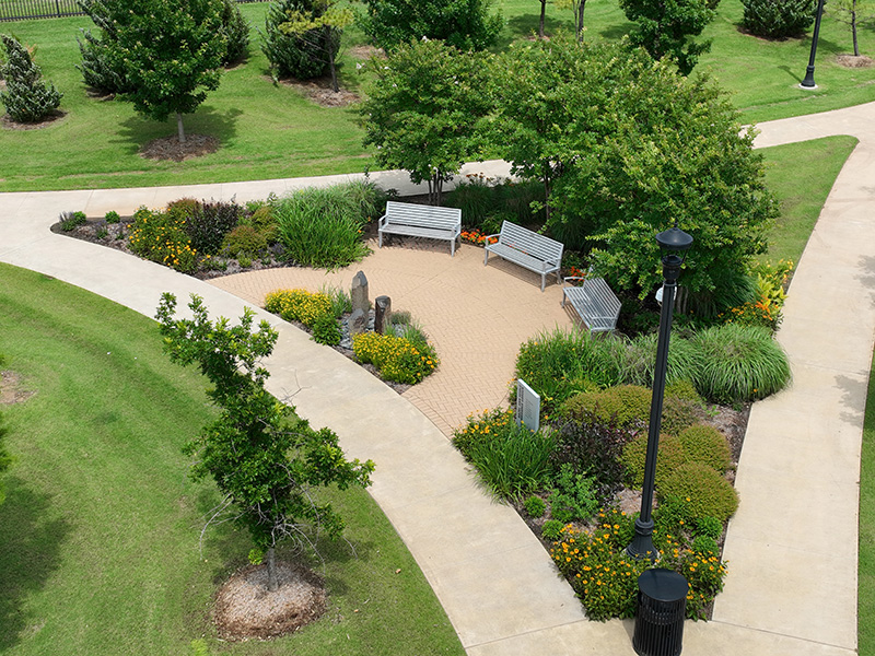 Aerial view of a park with a Y-shaped paved walkway surrounding a small garden area. The garden contains green plants, shrubs and yellow flowers. Two benches, a few stone markers and a black lamppost are within the garden. 