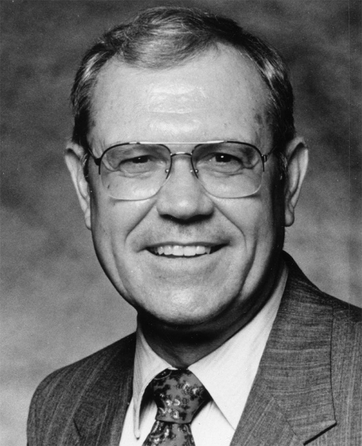 A grayscale portrait of a man wearing a suit jacket, dress shirt and patterned tie. The background is plain and out of focus.