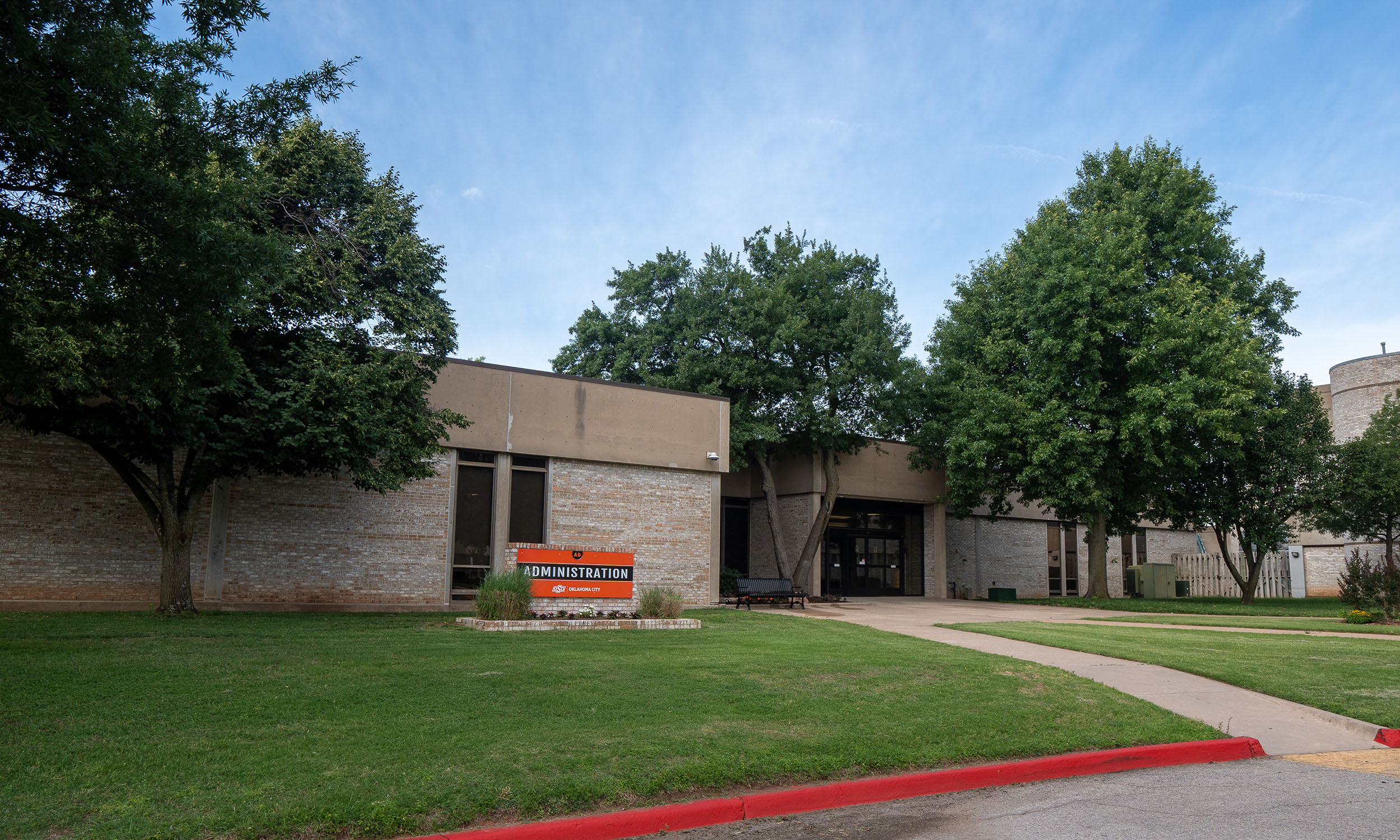 A single-story building with a flat roof, surrounded by trees and a well-maintained lawn. An orange sign in front of the building reads 'Administration.