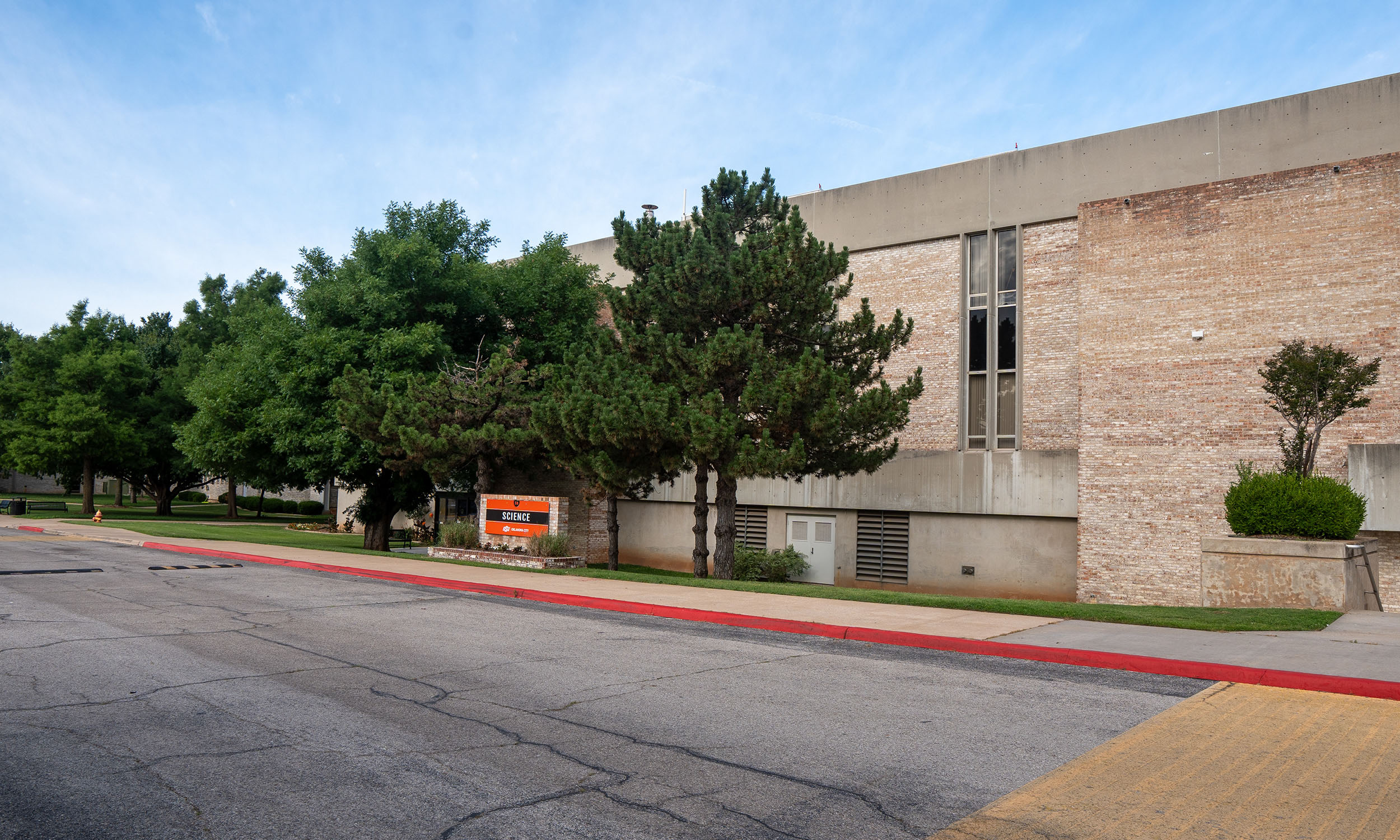 A large brick building with multiple windows and a 'Main Entrance' sign near the front. Trees and shrubs are landscaped in front, with a red curb lining the street. 