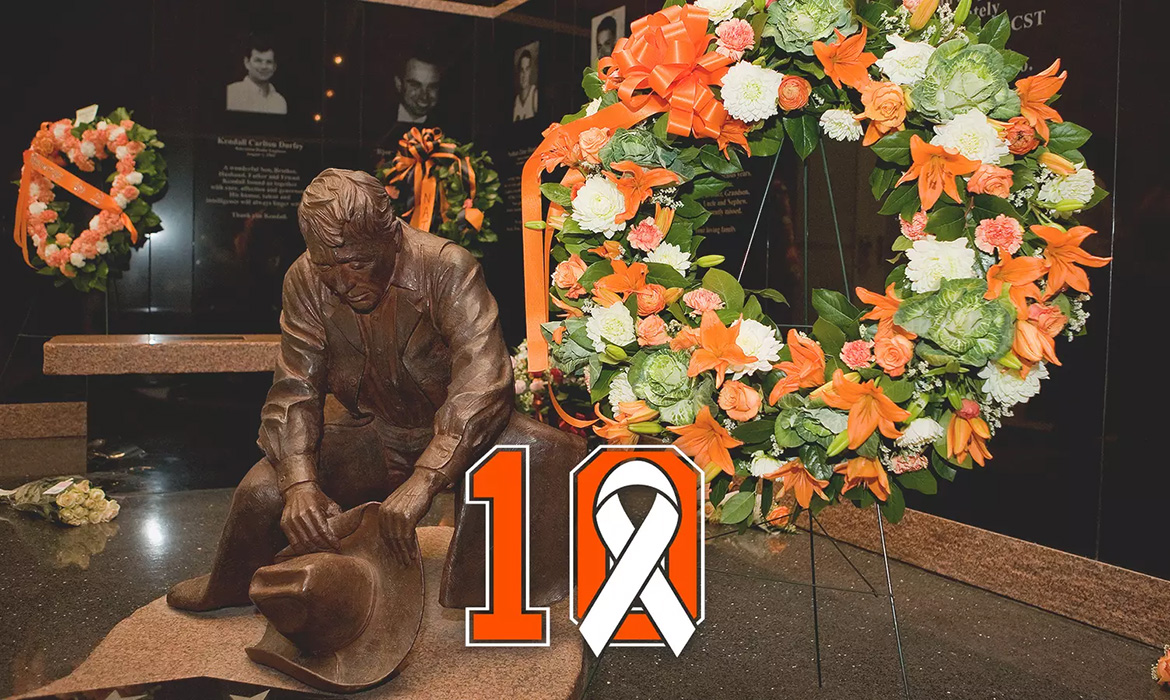 Bronze statue of a person kneeling and holding a hat, positioned in front of a memorial wall. Two large wreaths with orange and white flowers are placed on either side of the statue. The number '10' with an orange ribbon is prominently displayed in the foreground.